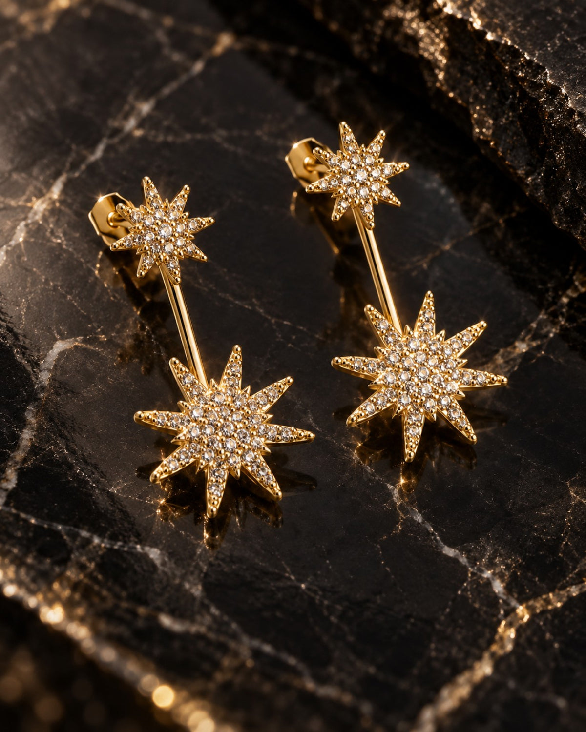 Par de brincos Celeste dois em um folheados a ouro, com duas estrelas cravejadas com zircônias em cada brinco, sobre pedra escura.