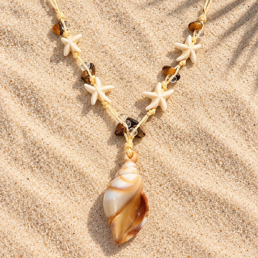 Colar Amara disposto sobre a areia, com pingente em formato de concha em tons de marfim e caramelo, estrelas-do-mar e contas marrons.