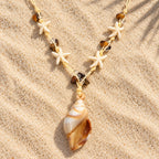 Colar Amara disposto sobre a areia, com pingente em formato de concha em tons de marfim e caramelo, estrelas-do-mar e contas marrons.