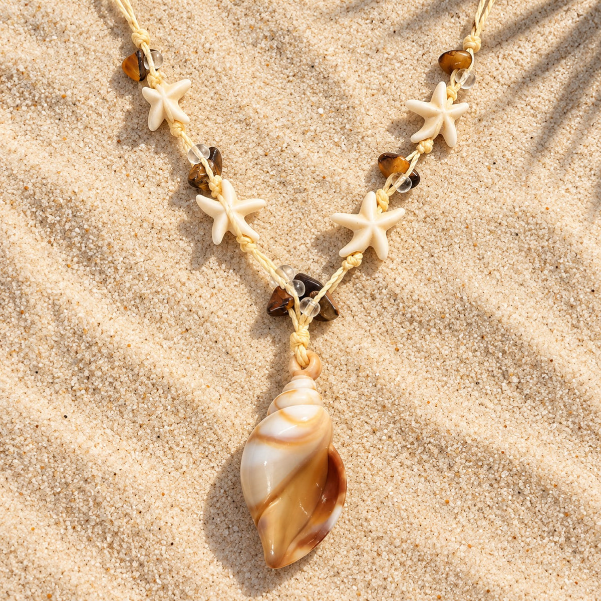 Colar Amara disposto sobre a areia, com pingente em formato de concha em tons de marfim e caramelo, estrelas-do-mar e contas marrons.
