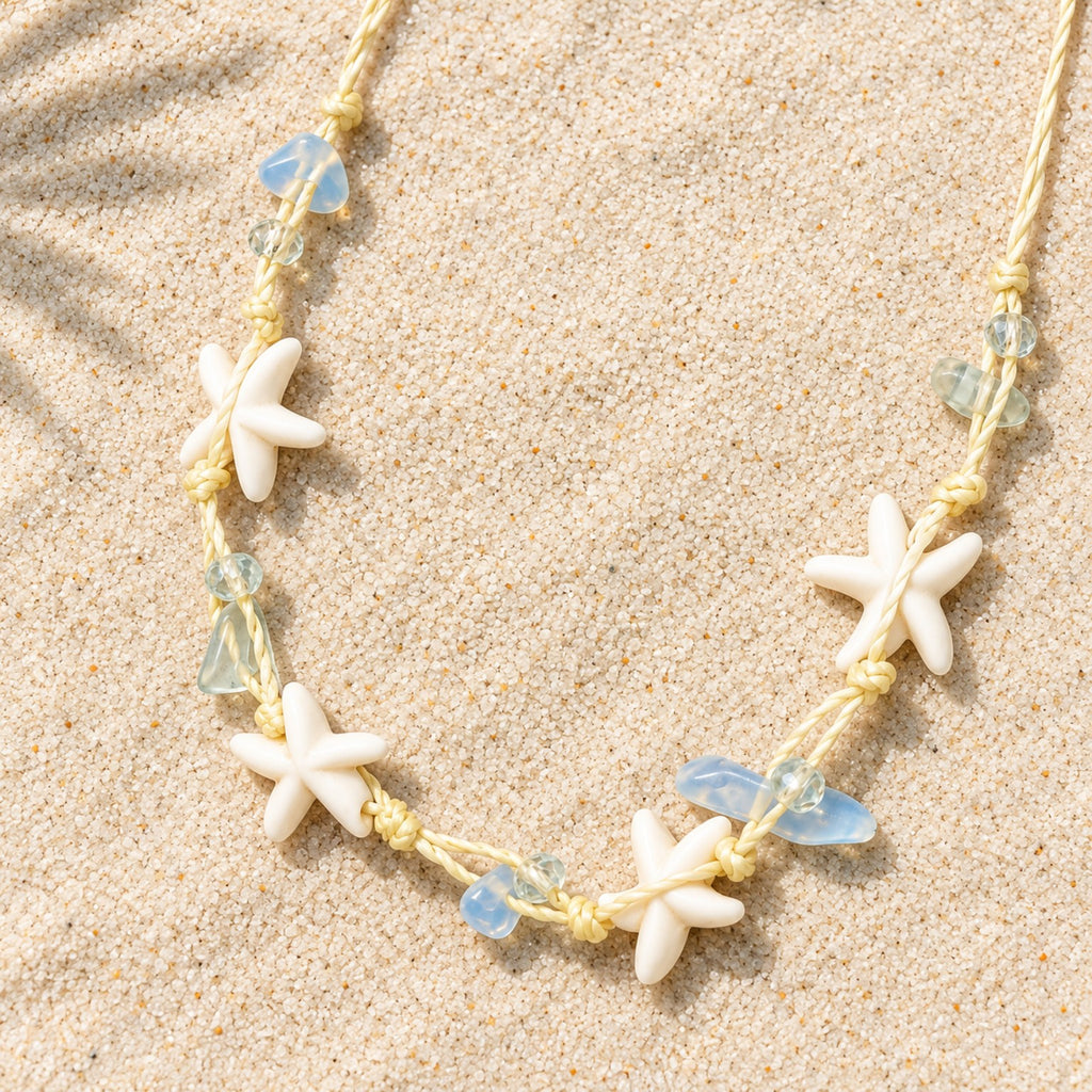 Colar Marina em material sintético, com estrelas-do-mar em tom marfim e contas azul-claras, disposto sobre a areia.