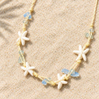 Colar Marina em material sintético, com estrelas-do-mar em tom marfim e contas azul-claras, disposto sobre a areia.