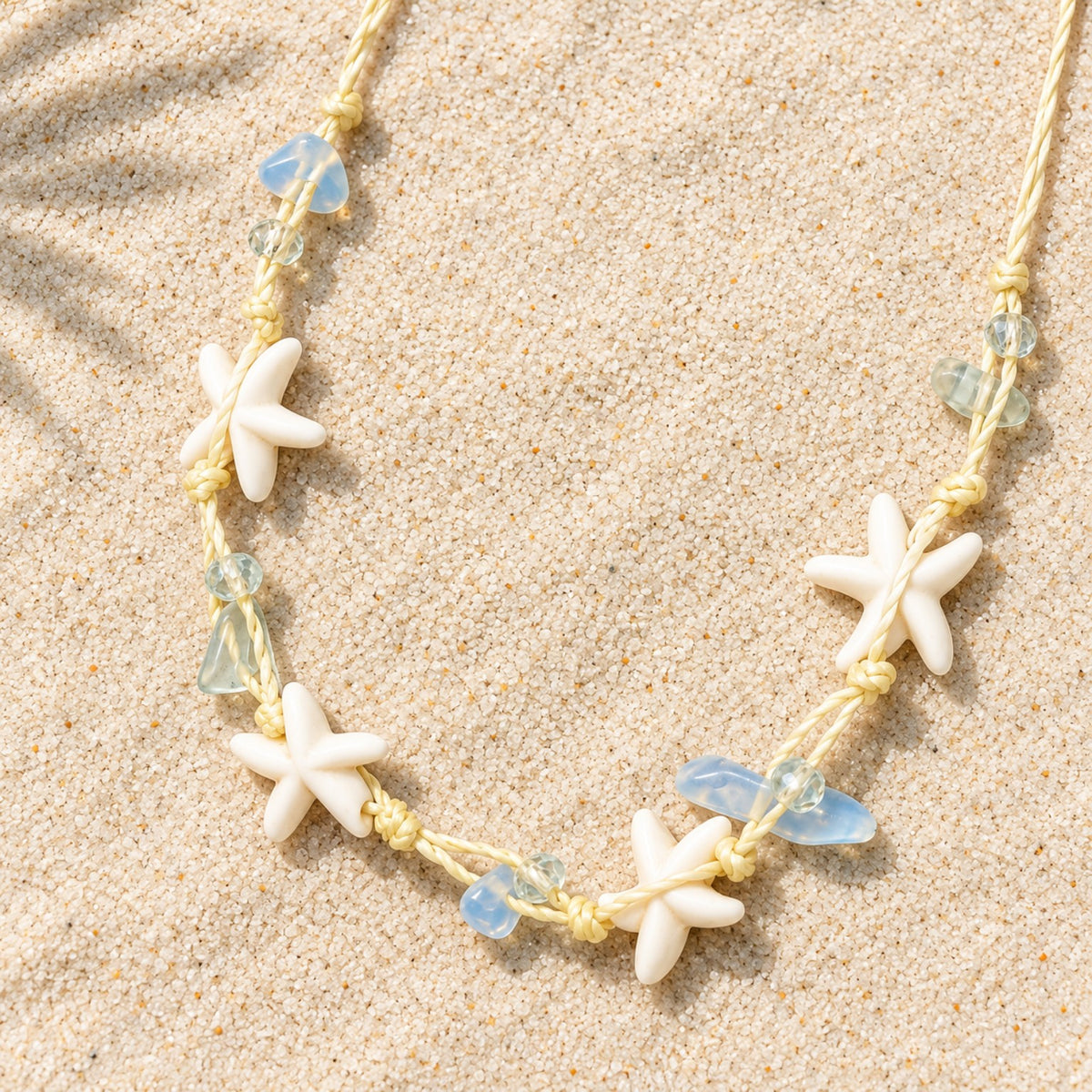 Colar Marina em material sintético, com estrelas-do-mar em tom marfim e contas azul-claras, disposto sobre a areia.
