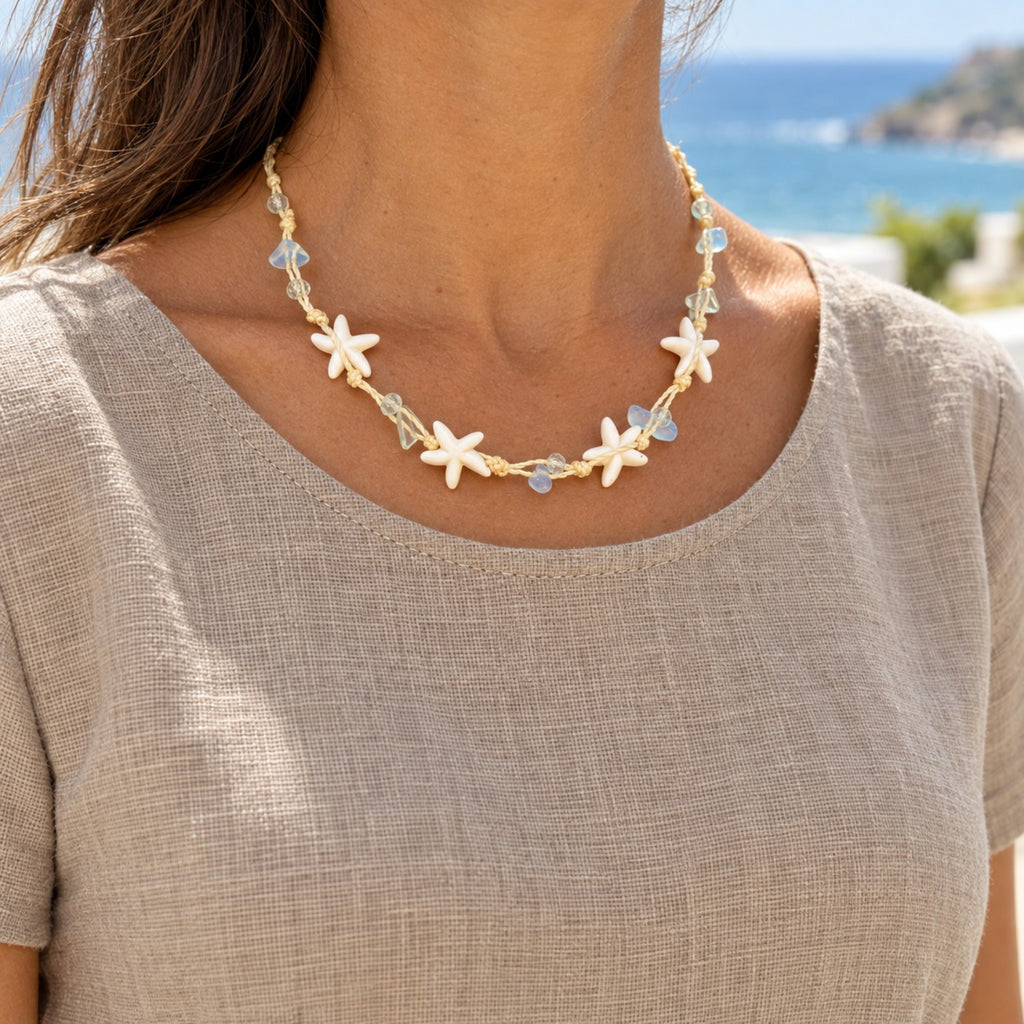 Modelo com blusa clara usando o Colar Marina Estrelas do Mar Azul, com o mar ao fundo.