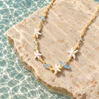 Colar Marina com estrelas-do-mar em tom marfim, contas azul-claras e cordão bege sobre uma pedra clara.