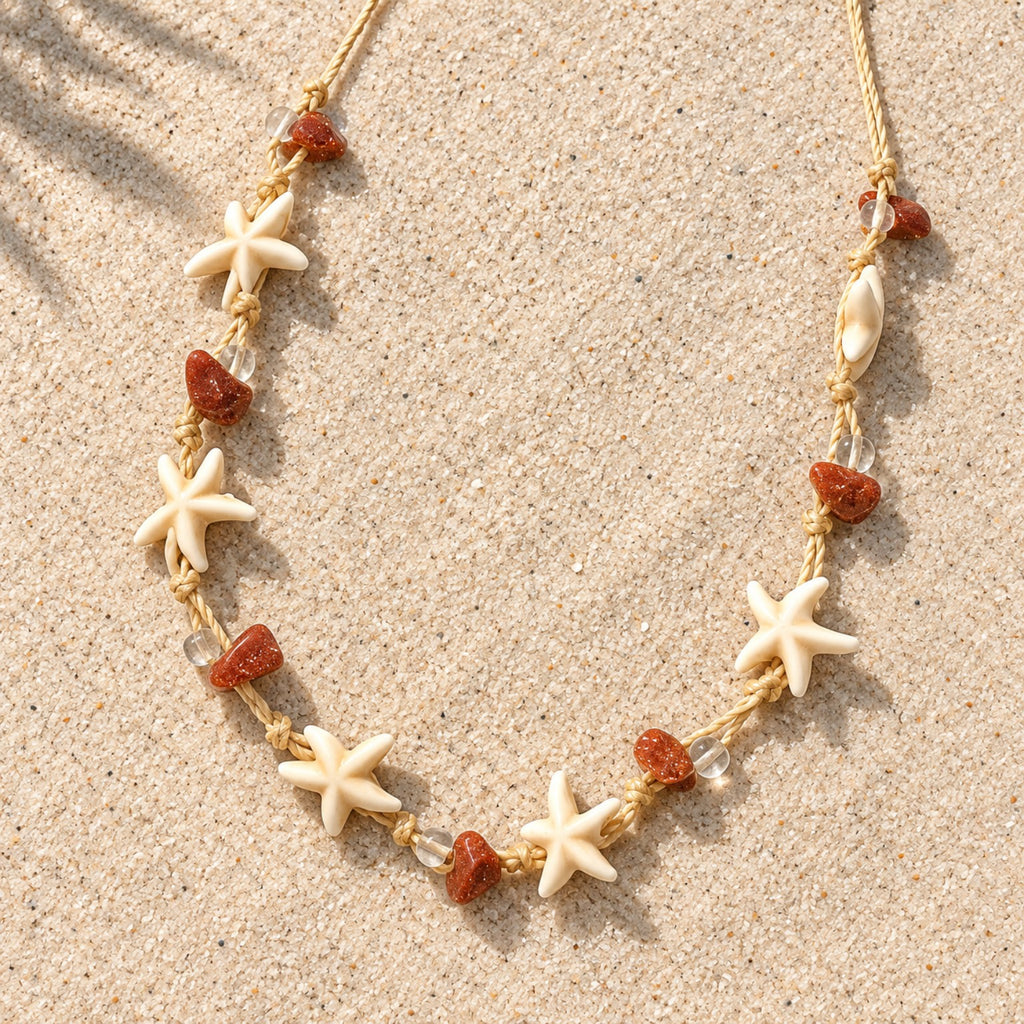 Colar Nalu disposto sobre a areia, com cordão bege, estrelas-do-mar em tom marfim e contas em tom terracota.