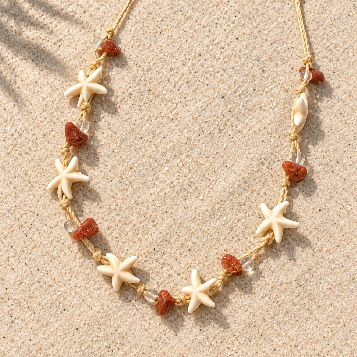 Colar Nalu disposto sobre a areia, com cordão bege, estrelas-do-mar em tom marfim e contas em tom terracota.