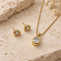 Conjunto Clara folheado a ouro, com colar de pingente em gota e par de brincos com zircônias incolores, sobre pedra clara.