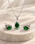 Conjunto Olívia em aço inoxidável prateado com colar e par de brincos em formato de gota verde, detalhes de zircônias e fundo de pedra clara.