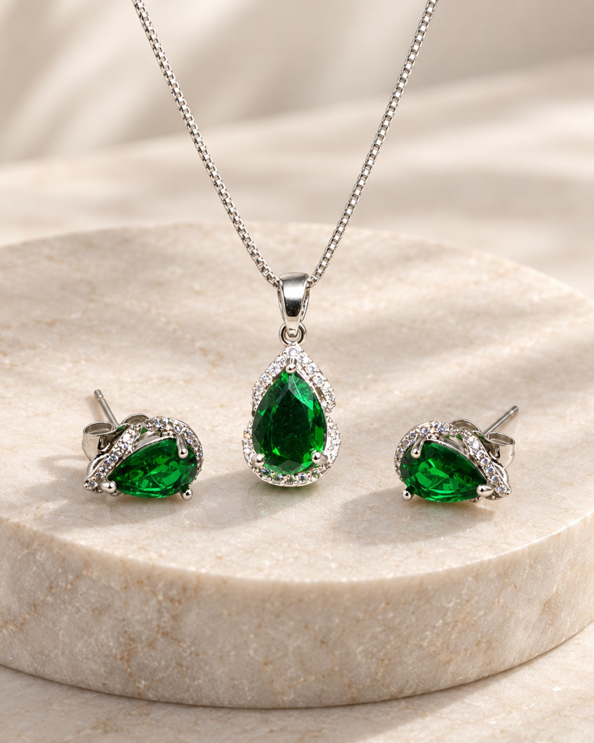 Conjunto Olívia em aço inoxidável prateado com colar e par de brincos em formato de gota verde, detalhes de zircônias e fundo de pedra clara.